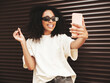 © halayalex - Beautiful woman with afro curls hairstyle.Smiling hipster model in white t-shirt. Sexy carefree female posing in the street near brown wall in sunglasses.Looking at smartphone screen, using apps