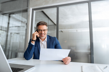 Positive financial specialist senior man working on laptop computer in office, having cell phone call. Mature businessman professional using pc, analysing business project, holding report. Copy space
