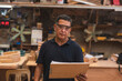 © Mdv Edwards - Portrait of a furniture workshop supervisor wearing safety glasses and holding a clipboard, standing in a busy carpentry environment.