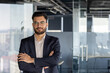 © Liubomir - A smiling businessman with arms crossed, standing in an office with a window background. He is wearing glasses and a suit.