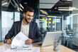 © Liubomir - A smiling businessman reviews documents, with a laptop open on his desk in a modern office setting, ready to work.