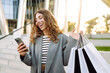© maxbelchenko - Online shopping! Happy woman with shopping bags with phone near shopping mall. Young woman with smartphone after shopping, enjoying sunset outdoors. Consumerism concept.