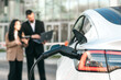 © wedmoments.stock - Charging electric car in the background of two business people with laptop. Concept of ecological transport and business.