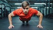 © vefimov - A young man in athletic clothing doing push-ups with a focused expression on his face, set against the backdrop of a gym.