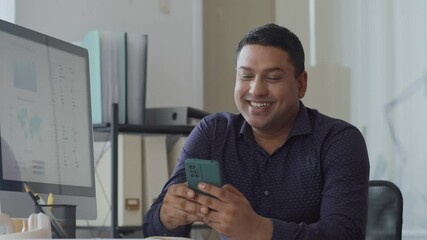 Medium shot of middle-aged Asian male employee feeling happy while texting on smartphone at office