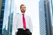 © Minerva Studio - Confident businessman standing with hands in pockets in front of skyscrapers
