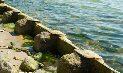 Fototapete - Series of weathered concrete blocks, showing signs of decay and algae growth, extend into the sea from a small, rocky beach