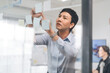 © dodotone - Business asian middle adult man using glass board adhesive note sticky ideas paper at indoors office