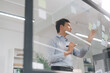 © dodotone - Business asian middle adult man using glass board adhesive note sticky ideas paper at indoors office