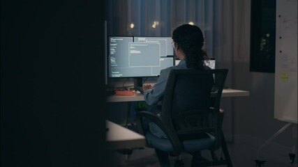 Back view of a female robotics engineer working late at night on multiple monitors, writing code and testing robotic systems in a dimly lit, modern tech workspace.