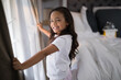 © wavebreak3 - Girl pulling beige curtains and smiling in bright bedroom, with white bed linens and morning light