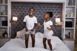 © wavebreak3 - African American child siblings jumping on bed in bedroom, with headboard pillows lamps shelves