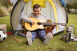 © wavebreak3 - child boy strumming acoustic guitar in backyard, with yellow-gray tent and camping gear