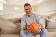 © wavebreak3 - Middle-aged man sitting on beige couch in living room, holding basketball with throw pillows
