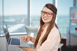© WavebreakMediaMicro - Young woman standing in office holding laptop and tapping trackpad, with eyeglasses and notes board