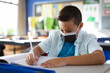 © Wavebreak Media - Elementary school boy writing in notebook at school desk, using white pencil and wearing face mask