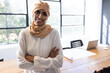 © Wavebreak Media - Mid adult Indian woman standing with arms crossed in design studio, with laptop and blueprints
