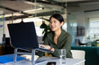 © Wavebreak Media - Asian woman typing on laptop on stand at open-plan office desk with smartphone, folder, water glass