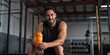 © Usman - Man sitting in gym holding orange shaker bottle after workout with a smile on his face looking forward