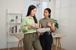 © Johnstocker - Portrait of Two Asian businesswomen stand in an office, one holding a document explaining a job, while the other listens intently and sips coffee.