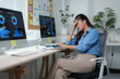© crizzystudio - Young businesswoman sitting at her desk, experiencing headaches and work-related stress while analyzing graphs and charts on computer screens in a modern office