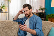 © Andrii Iemelianenko - Middle-aged man at home feeling frustrated and nervous while texting on smartphone, holding head in distress. Nervous Caucasian guy sitting on sofa, anxiously waiting for an important message reply
