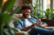 © Aisylu - Handsome man relaxing on comfy armchair among home plants holding in hands smartphone. No stress, home owner, modern tech usage, fun concept. AI