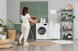 © Pixel-Shot - Young woman with wicker basket walking towards modern washing machine in laundry room