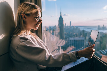 Wall Mural - Happy female blogger watching positive video vlog on streams website connecting to 4g internet on laptop computer, cheerful hipster girl networking near panoramic window with metropolitan scenery