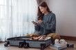 © Tj - Happy asian woman packing clothes into travel bag, using mobile phone, preparing for vacation trip, sitting near window at home, booking hotel or tickets online