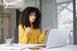 © Liubomir - A woman with curly hair appears frustrated during a video call, gesturing with her hands while seated in front of a laptop.