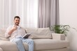 © New Africa - Happy man drinking coffee while reading book on sofa near window with beautiful curtains at home in living room. Space for text