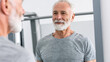 © Athiphat - senior man with gray beard smiles at his reflection in fitness mirror, showcasing positive attitude towards health and wellness. setting emphasizes modern gym environment