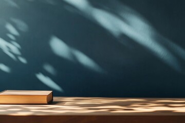Poster - A closed hardcover book rests on a wooden desk, radiating a gentle glow from its pages while soft shadows of leaves create a whimsical ambiance on the wall