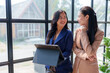 © crizzystudio - Two asian businesswomen are standing near a large window in a modern office, engaged in a discussion while one of them holds a digital tablet, possibly reviewing data or strategizing