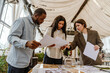 © Drobot Dean - A young Black groom and bride are discussing printed materials with a young White female wedding planner in a decorated event venue filled with natural light and draped fabric ceiling.