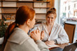 © Drobot Dean - Two White women with red hair in their 20s sit at a wooden table with a laptop and a notebook in a cafe talking, one in a white shirt gesturing with a pen, the other wearing a grey sweater listening.