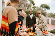 © Drobot Dean - A multiethnic group of casually dressed smiling friends, including White and Black in their 20s and 40s, stand outdoors on a patio around a table full of food, talking, eating food, and drinking wine.