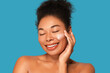 © Beauty Agent Studio - Radiant African American woman with curly hair in a high bun, smiling with closed eyes while gently applying skincare hydrating  cream to her cheek against the bright turquoise background.