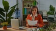 © kinomaster - Older adult businesswoman works at green office with plants, woman sitting at desk working on laptop, checking looking at paper documents.