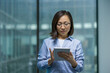 © Liubomir - Serious and confident office worker using tablet computer. Asian businesswoman using online financial app, reading thoughtfully while standing near window inside office.