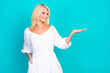© deagreez - Blonde senior woman in a white summer dress gesturing with her hand against a teal background