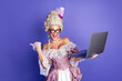 © deagreez - Young female in historical aristocratic attire holding a laptop, set against a vibrant purple background
