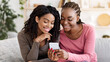 © Prostock-studio - Smiling african american girlfriends checking photos on mobile phone, spending time together at home, copy space. Two young black ladies sitting on sofa at living room, using modern smartphone
