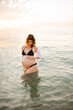 © morrowlight - Pregnant woman wearing black swimsuit in sea water over sunset outdoors. Summer vacation time. Maternity.