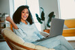 © deagreez - Young smiling woman with curly hair using a laptop in a cozy living room on a sunny day embracing modern technology and comfort