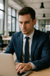 © JulieMeneghin - Jeune homme en costume travaillant sur un ordinateur portable dans un bureau moderne, concentré sur des tâches administratives ou financières dans un environnement lumineux