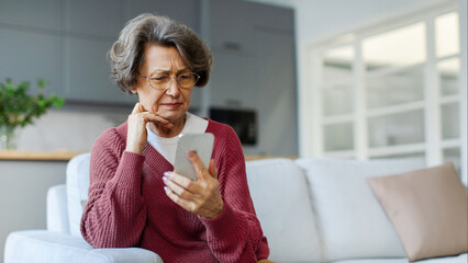 What should I press next. Thoughtful senior woman sitting on sofa holding cellphone and looking pensive on screen, texting message, browsing internet online, panorama, free space