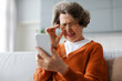 © Home-stock - Senior woman touching eyeglasses and squinting eyes, trying to read message on cellphone, sitting on couch at home