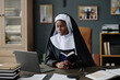 © AnnaStills - Medium shot of young adult African American nun holding bible in two hands while sitting in brown leather office chair, she looking at laptops screen while having video call